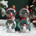 Four small snow globes with Christmas-themed figures on a snowy surface.