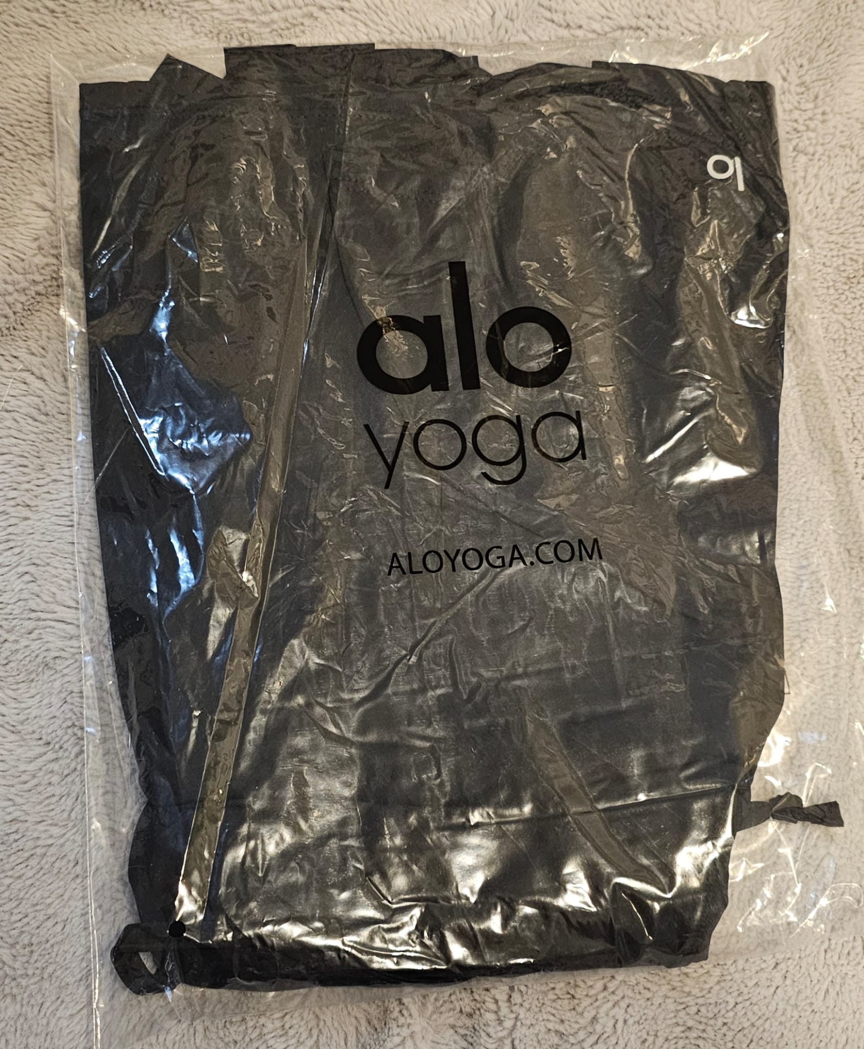 Black yoga mat in a clear plastic bag with 'alo yoga' branding on a textured surface.